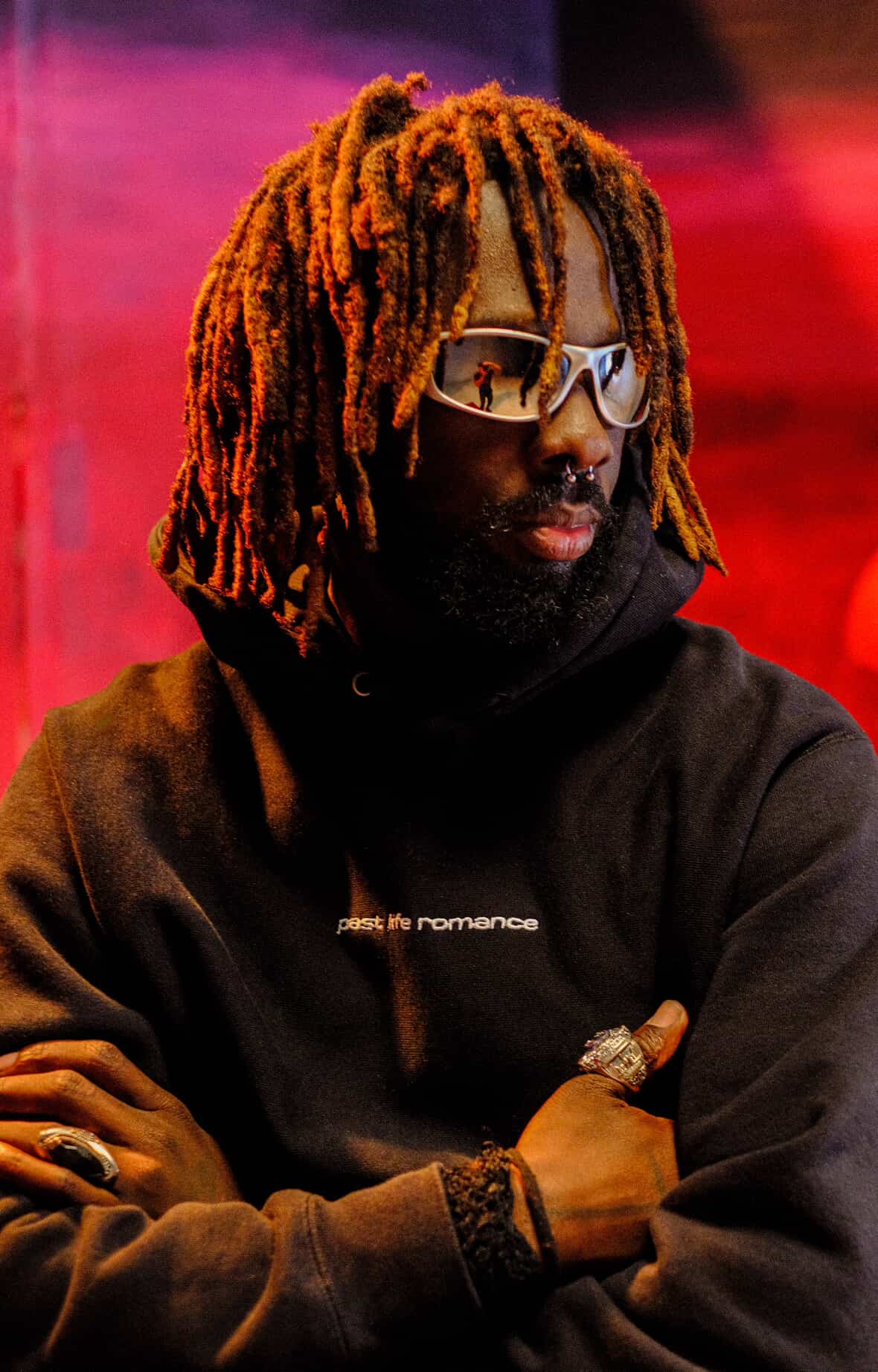 Person wearing a black hoodie with text, sunglasses, and multiple rings against a red background