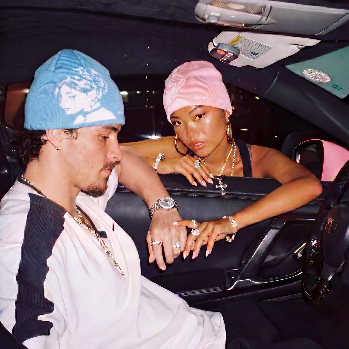 Two people sitting inside a car, one wearing a blue beanie and the other a pink headscarf.